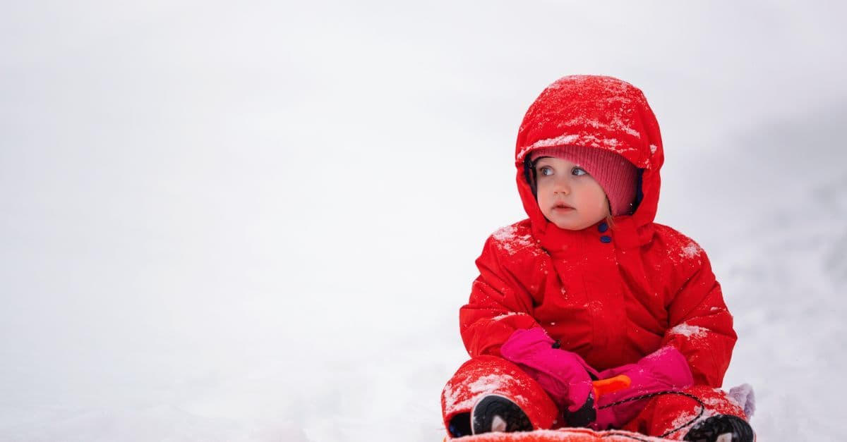 Kleines Kind im roten Schneeanzug sitzt auf einem Schlitten im Tiefschnee und schaut nach rechts.