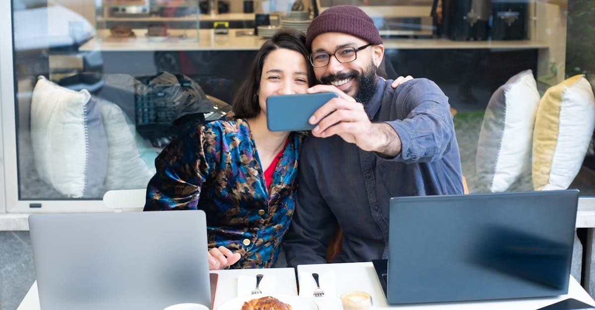 Pareja sonriente tomándose selfie en café con laptops y pasteles, conectados sin datos en su destino