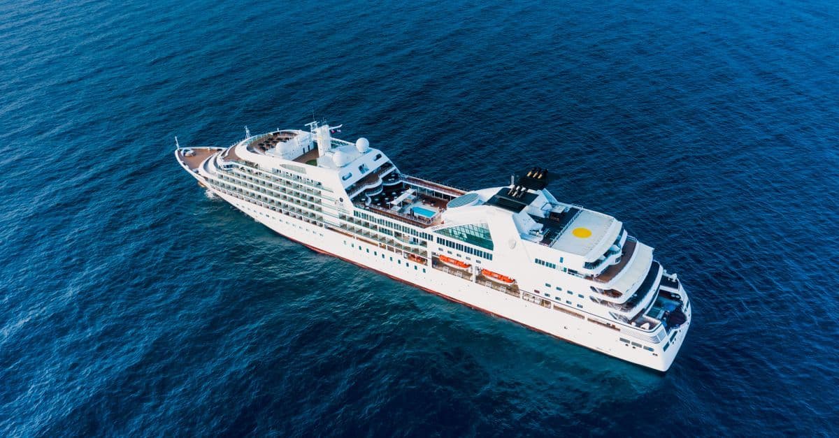 A large white cruise ship sails across deep blue open sea, seen from above.