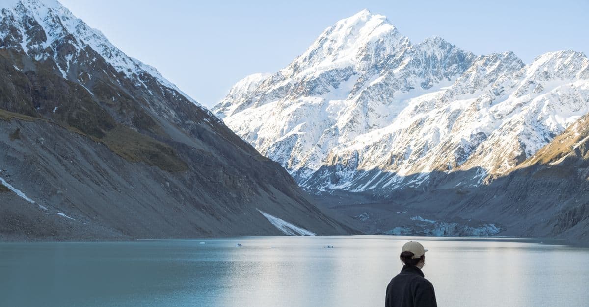 A traveler looks across a glacial lake toward snow-covered mountains in New Zealand.