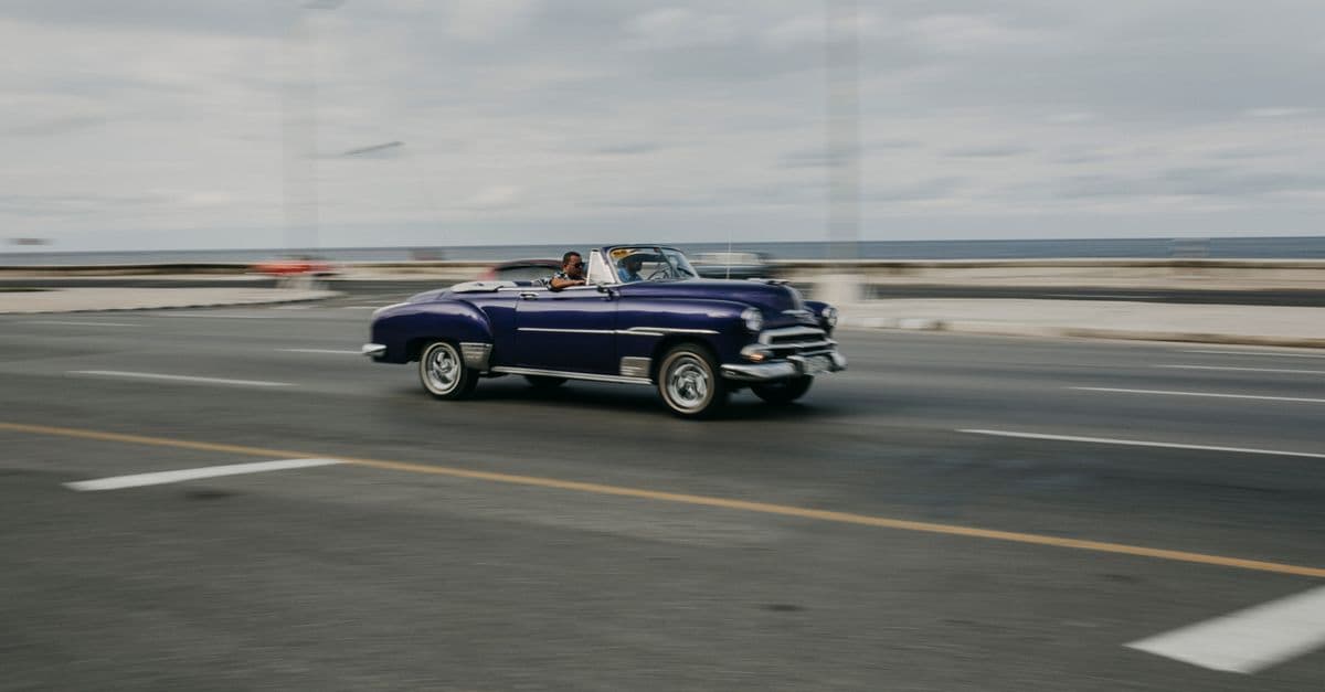 A classic blue convertible drives along a coastal road beside the sea.
