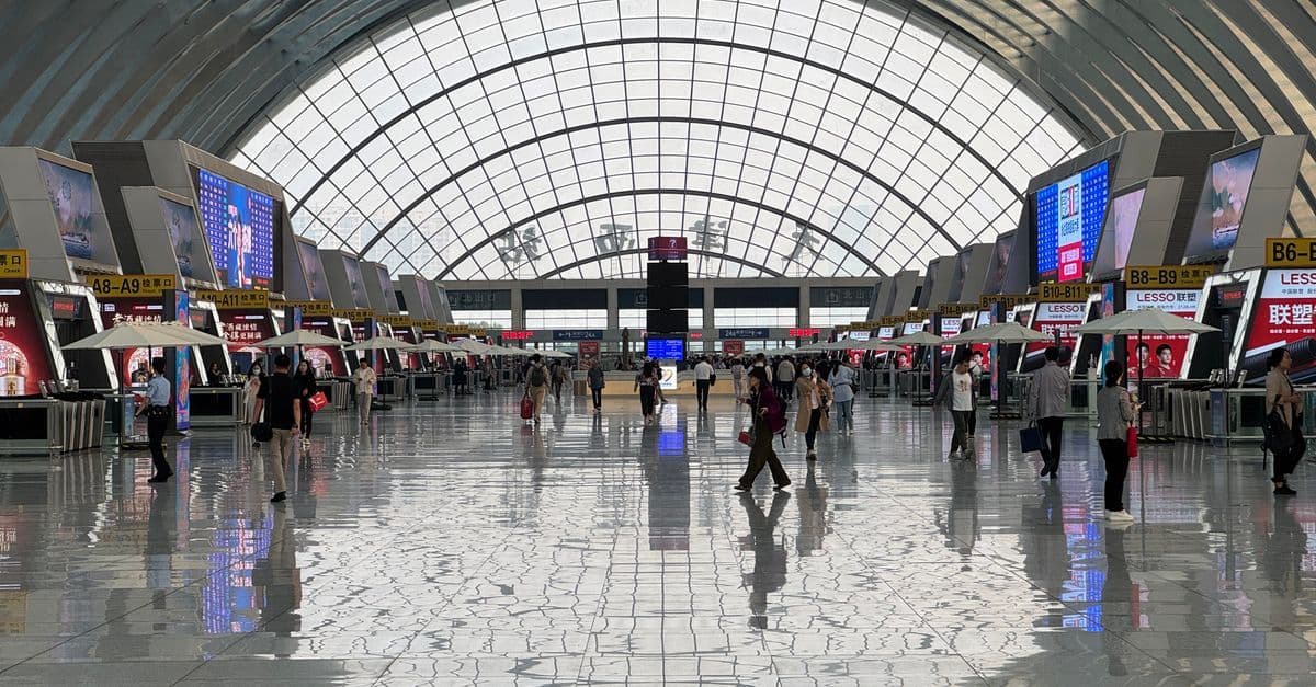 Travelers walk through a large airport terminal beneath a curved glass roof, with check-in counters along both sides.