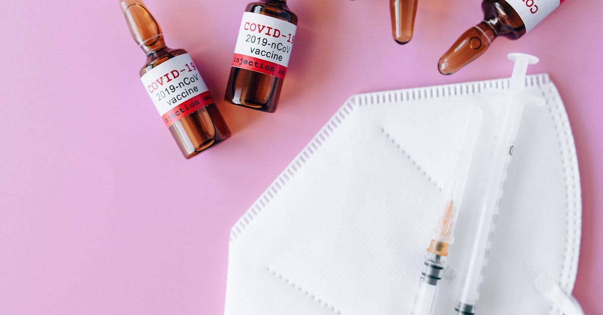 Flacons de vaccins, seringue et masque sur surface rose, rappelant l'importance du calendrier vaccinal avant le départ.