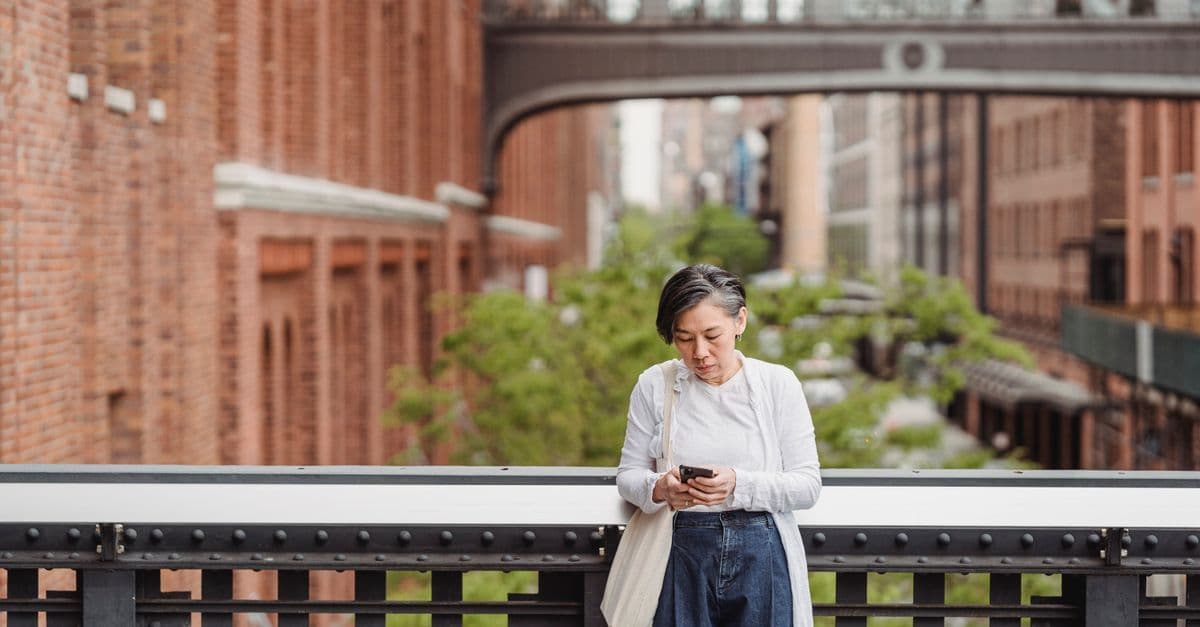 A woman looks down at her phone while leaning on a railing above a city street, a cloth tote on her shoulder.