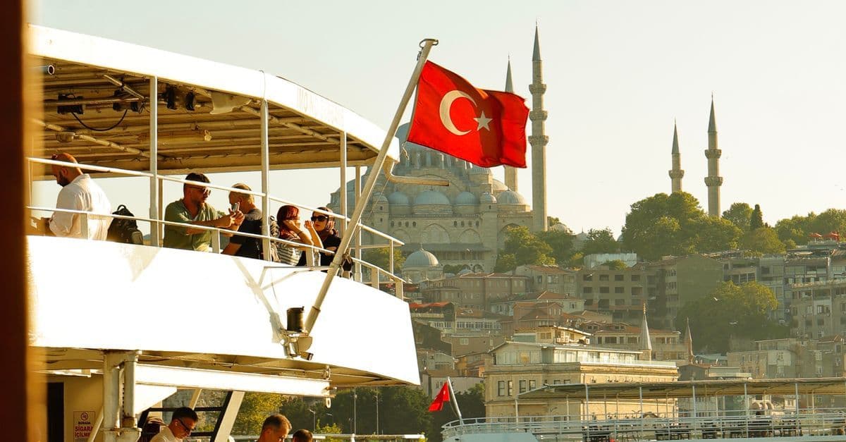 Fahrgäste auf dem Oberdeck eines Schiffs mit türkischer Flagge, dahinter eine große Moschee auf einem Hügel in Istanbul.