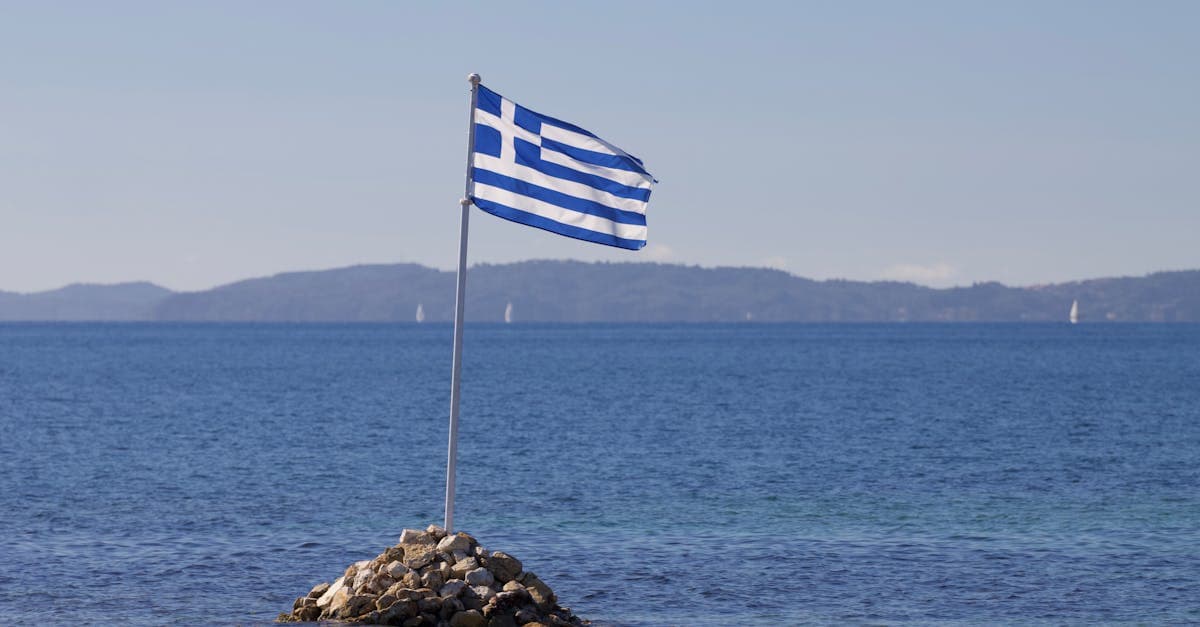 Grekisk flagga vajar över klippig kust på Korfu med det stilla Joniska havet i bakgrunden.