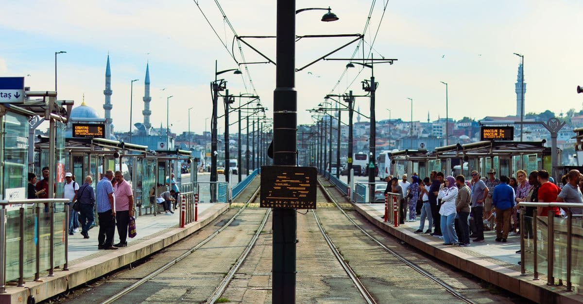 Fahrgäste warten an einer Straßenbahnhaltestelle in Istanbul, links ragen die Minarette einer Moschee in den Himmel.