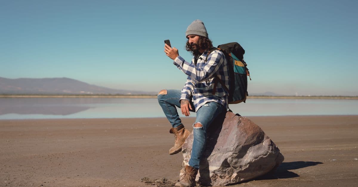 A backpacker sits on a rock by a calm lake and holds up his phone, with low hills on the far shore.
