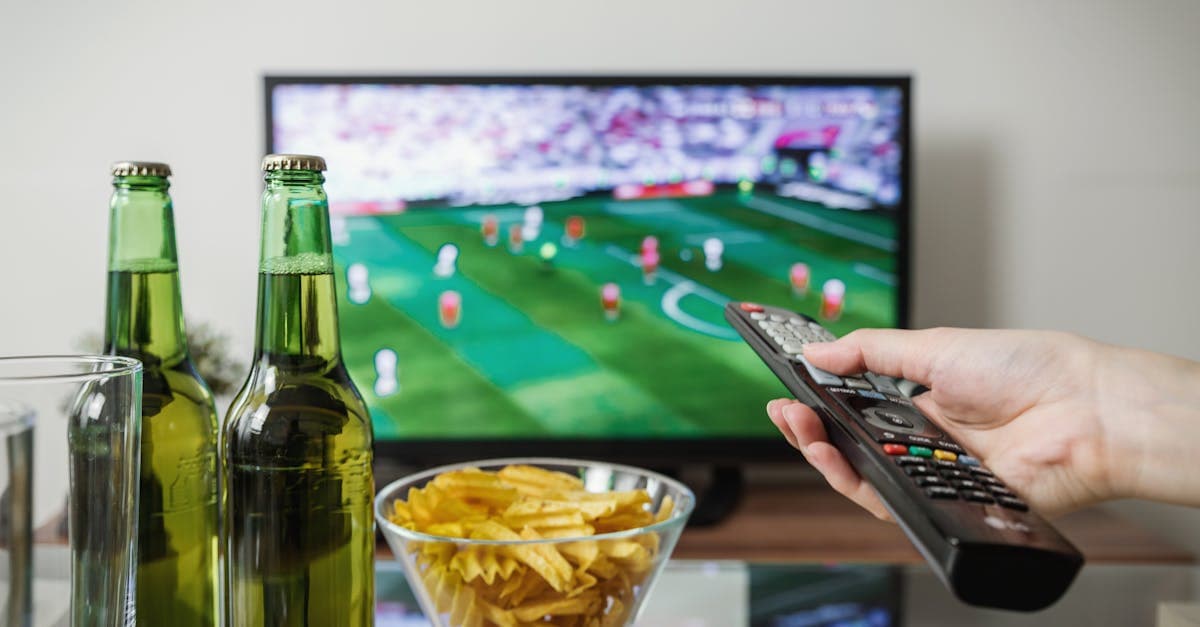 Person holding a remote watching soccer on TV with beer and snacks, streaming via streameast app.![image