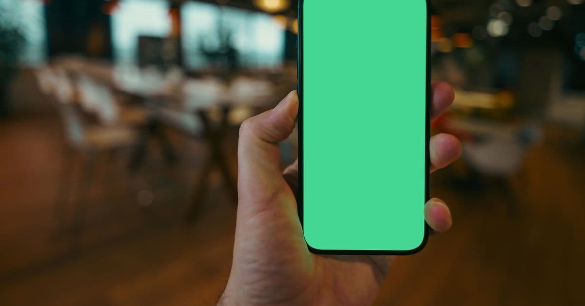 Hand holding a smartphone with a bright green blank screen against a blurred cafe interior background.