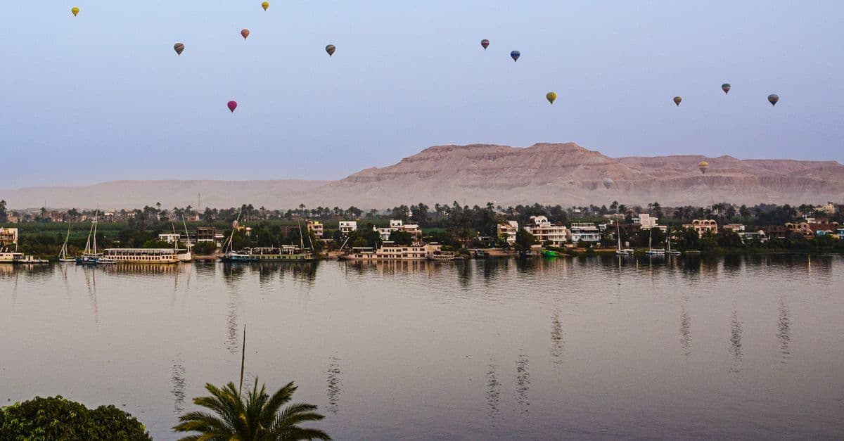 Hot air balloons float above the Nile and riverside homes, with desert mountains beyond.