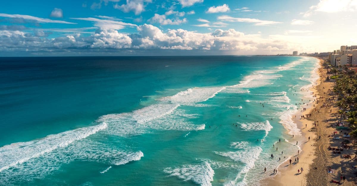 Turquoise waves roll onto Cancun’s sandy beach, lined with resort buildings and beachgoers.