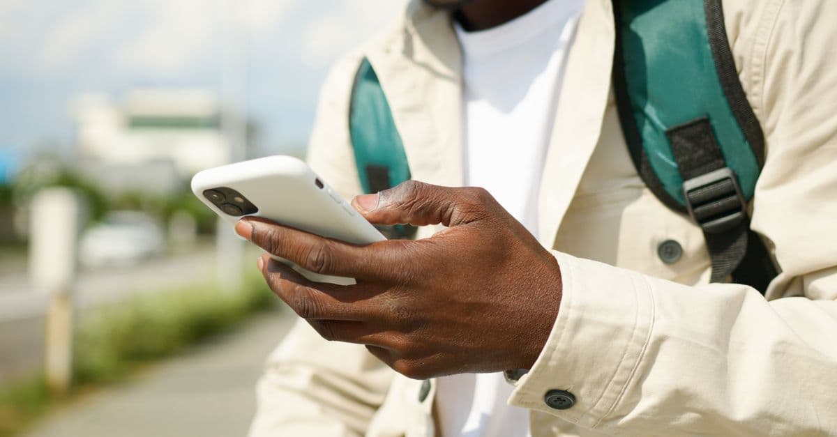 A traveler with a backpack strap over a light jacket holds a white smartphone outdoors.