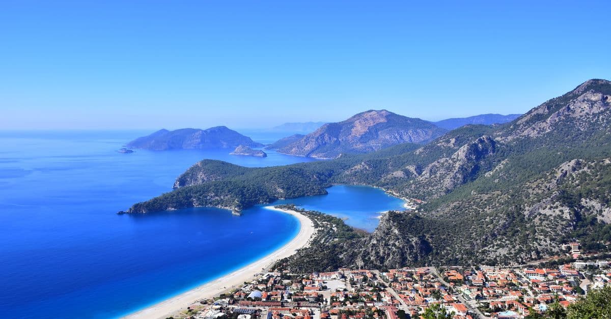 Widok z góry na zatokę Ölüdeniz w Turcji: długa plaża, turkusowa laguna i miasteczko u stóp gór.