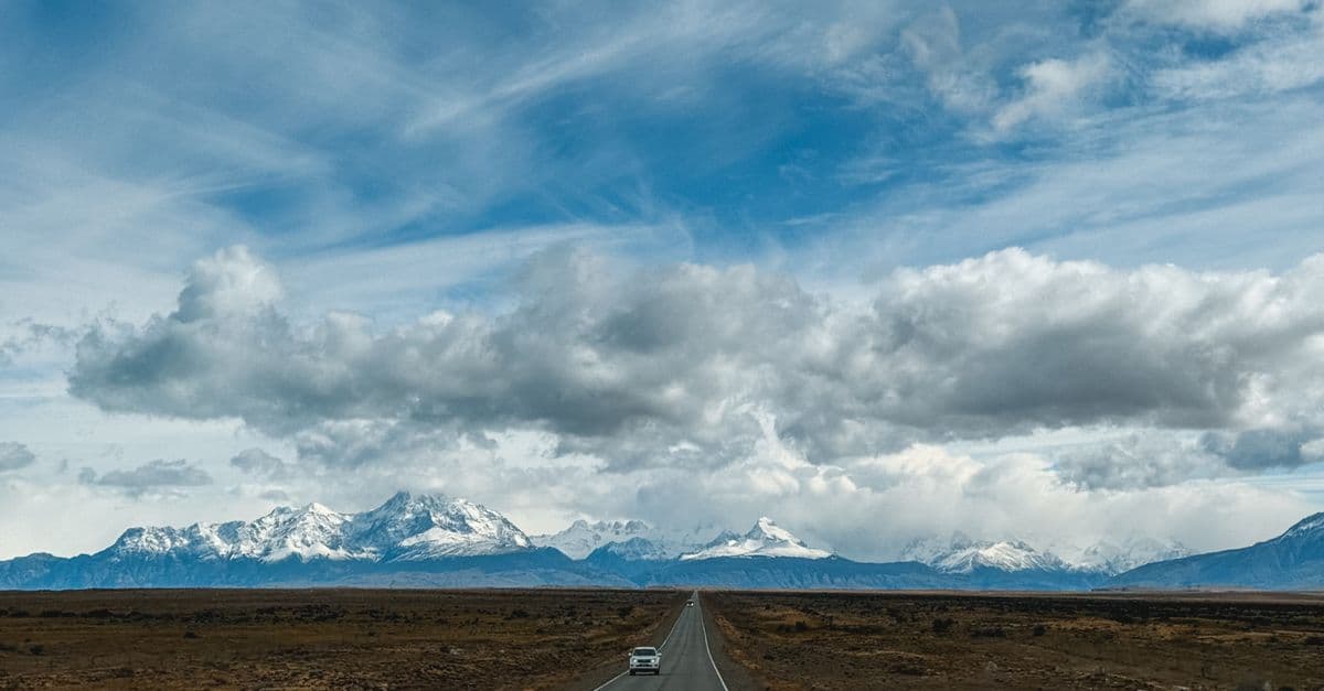 A lone car travels along a straight road across Patagonia's open plains toward snow-capped mountains beneath a cloudy sky.