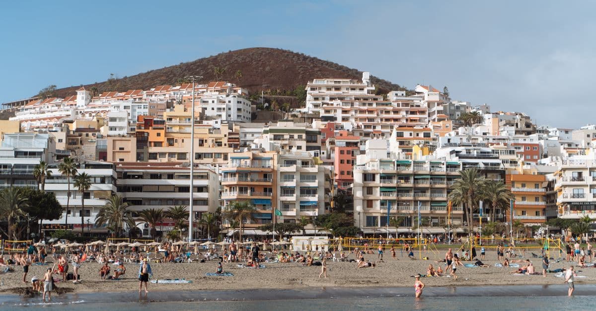 Sunbathers on a sandy beach below apartment blocks and a dry hill in Tenerife.