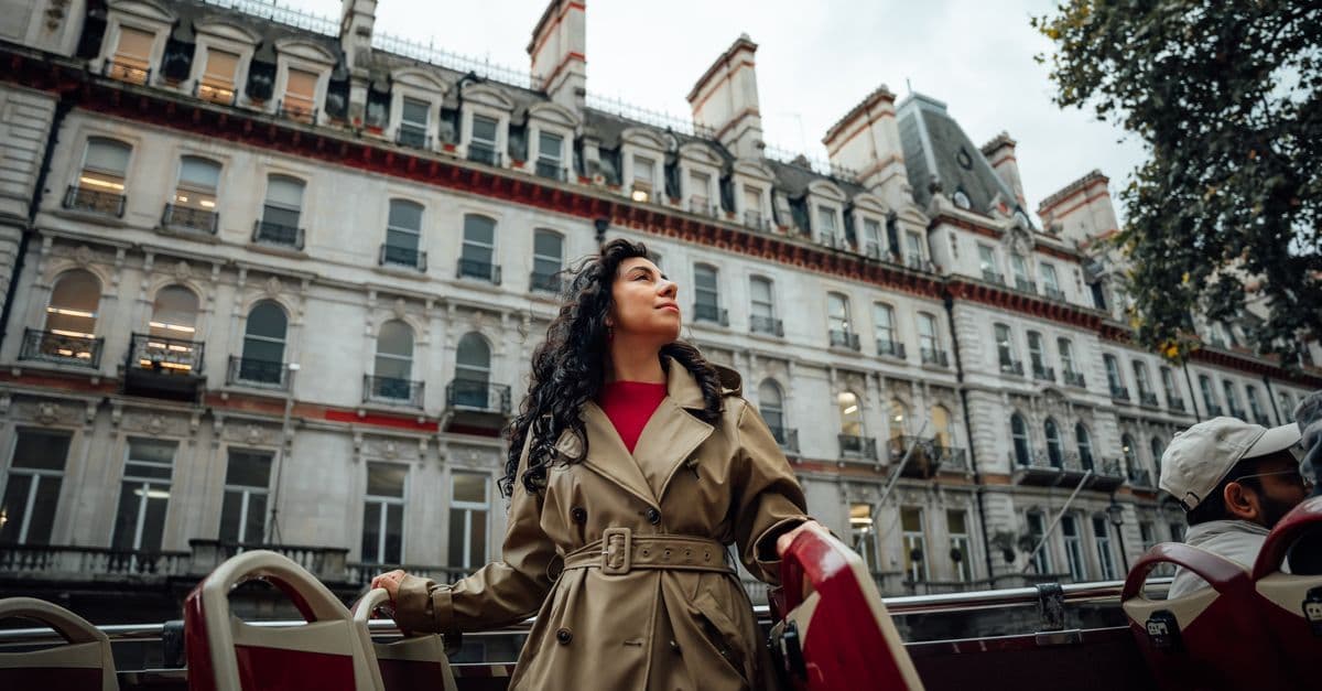 Mujer con gabardina disfruta de un autobús turístico descubierto rodeada de arquitectura histórica bajo el tiempo en Londres.