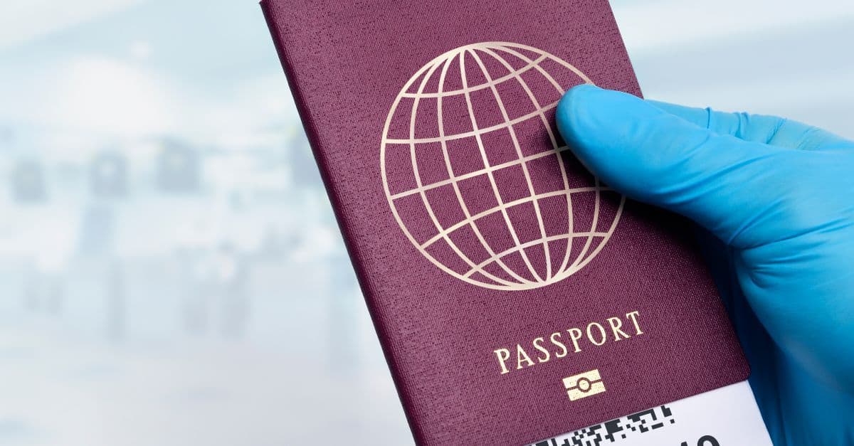 A blue-gloved hand holds a maroon passport with a globe emblem against a blurred background.