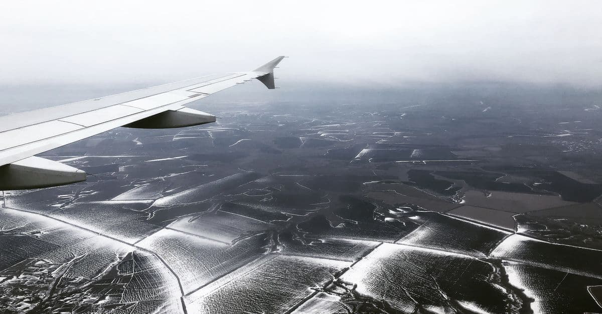 An airplane wing overlooks snow-dusted fields and dark countryside beneath a cloudy sky.
