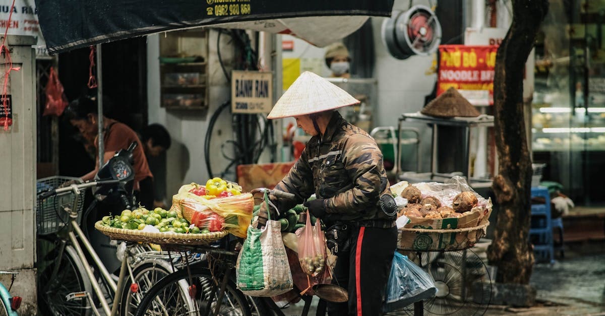 Traditional-hatted Hanoi market vendor selling fresh vegetables and fruits at a busy outdoor stall