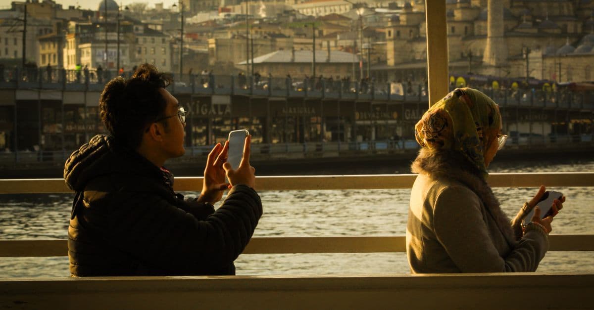 Two people use their phones by the water in Istanbul, with a busy bridge and old domes behind them.