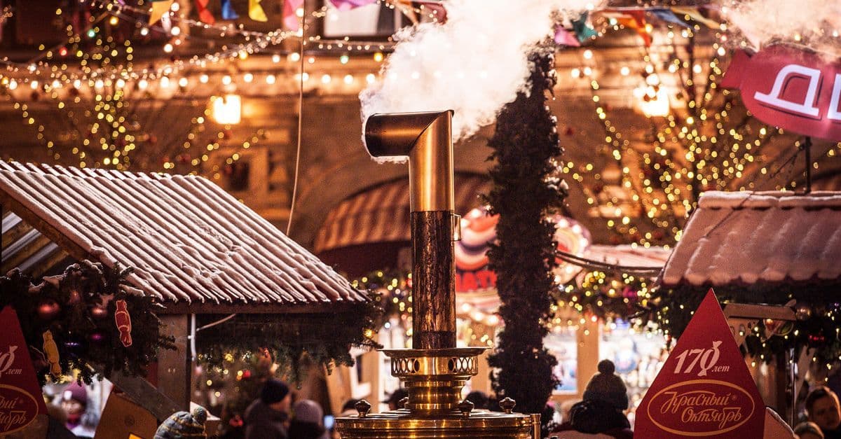 A steaming outdoor heater stands among warmly lit Christmas market stalls and shoppers at dusk.