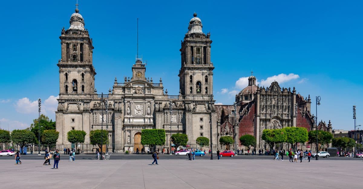 Mexico City’s Metropolitan Cathedral stands beneath a clear blue sky, with people crossing the open plaza.
