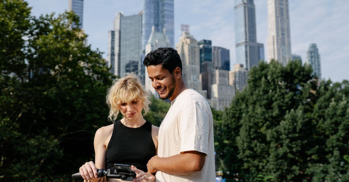 A couple smiles at a phone on a selfie stick in Central Park, New York, with tall towers rising behind the trees.