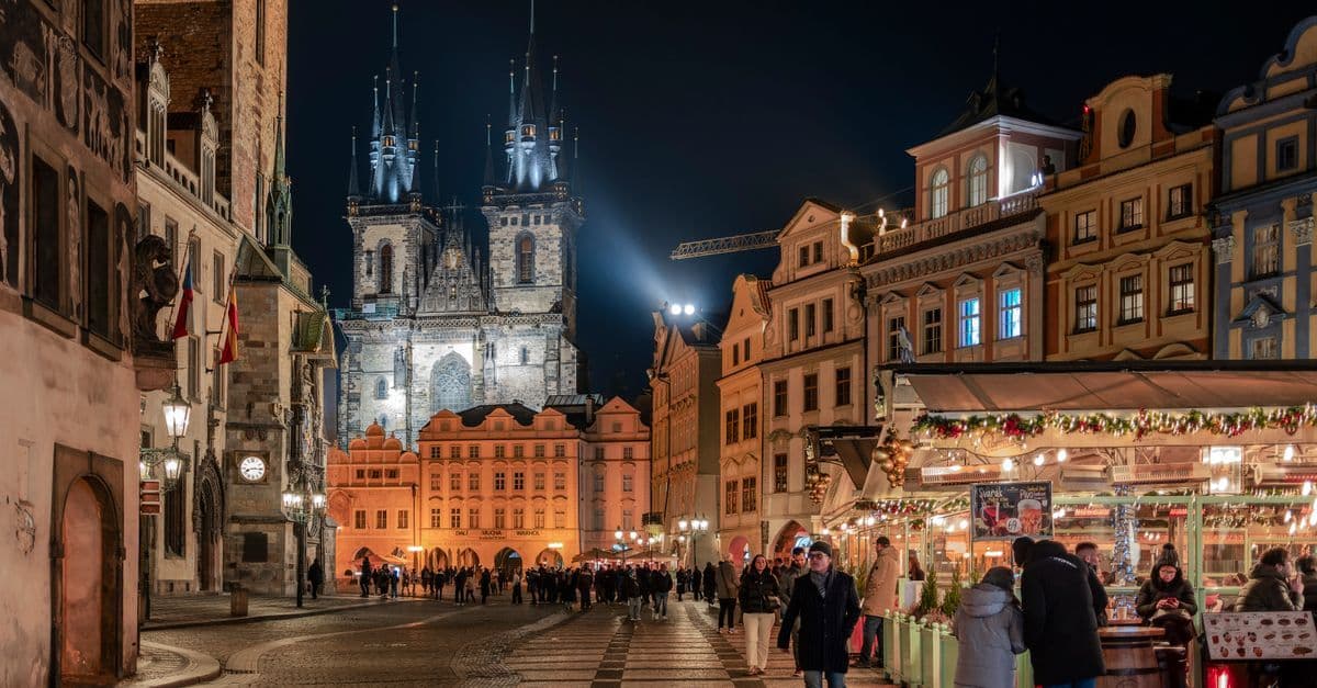 A brightly lit Christmas market stall fills Prague’s Old Town Square beneath the illuminated spires of a Gothic church.