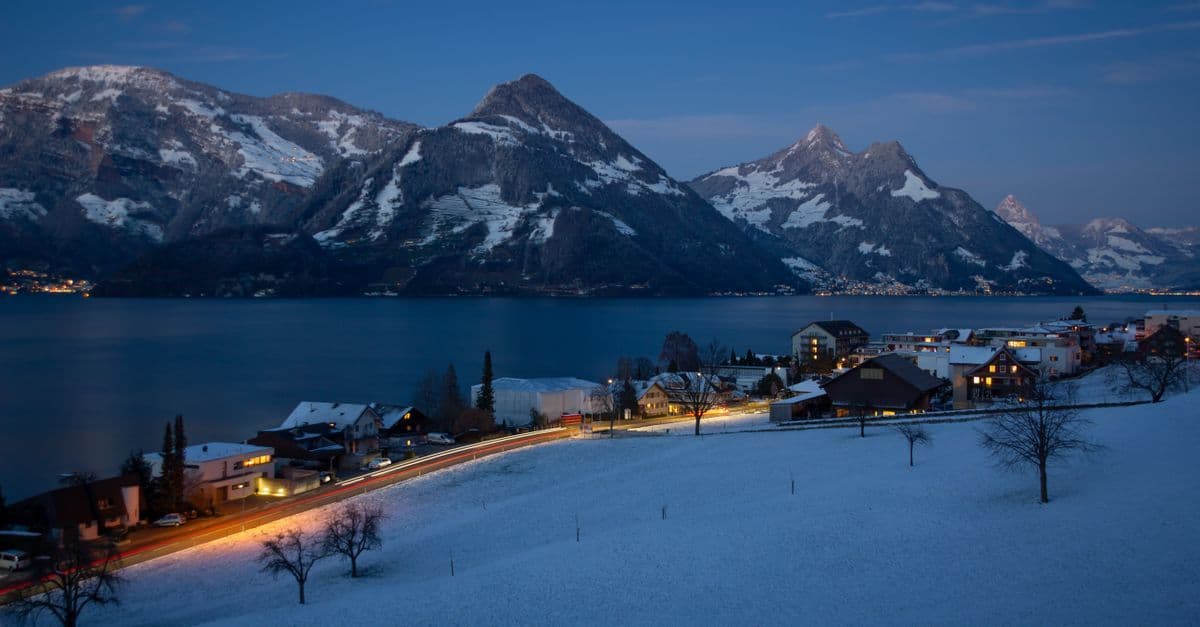 Verschneites Beckenried in der Abenddämmerung mit erleuchteten Häusern und Blick auf die Schweizer Alpen.