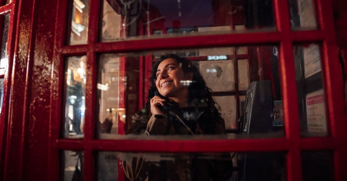 Woman smiling inside a red London phone booth at night, considering UK phone costs abroad.