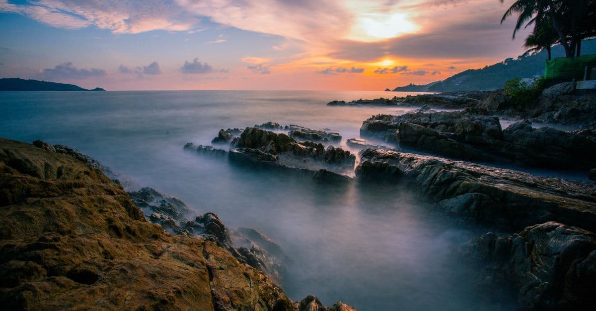A vivid sunset glows over the sea as waves wash around a rocky coastline lined with palm trees.