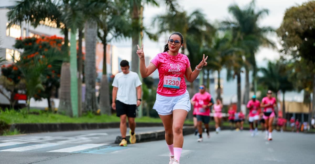 ヤシの木が並ぶ道路を、ピンクのランニングシャツと白いパンツ姿の女性がゼッケンを着けて笑顔で走り、後ろにも参加者が続いています。