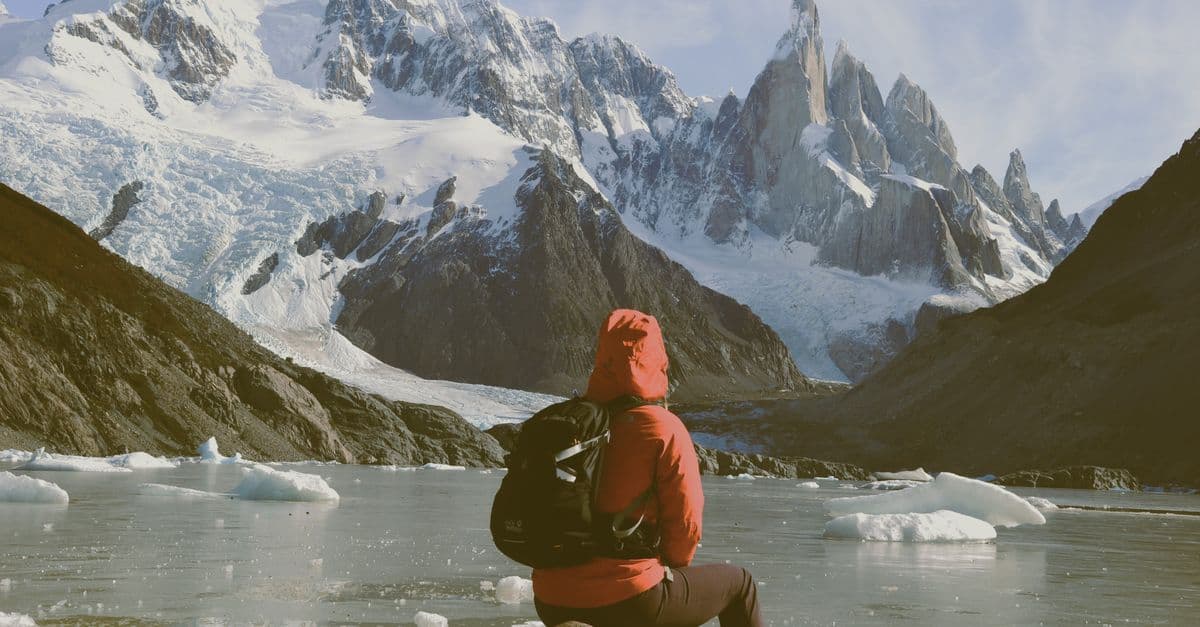A hiker in a red jacket sits beside an icy lake beneath the snow-covered peaks of Patagonia.