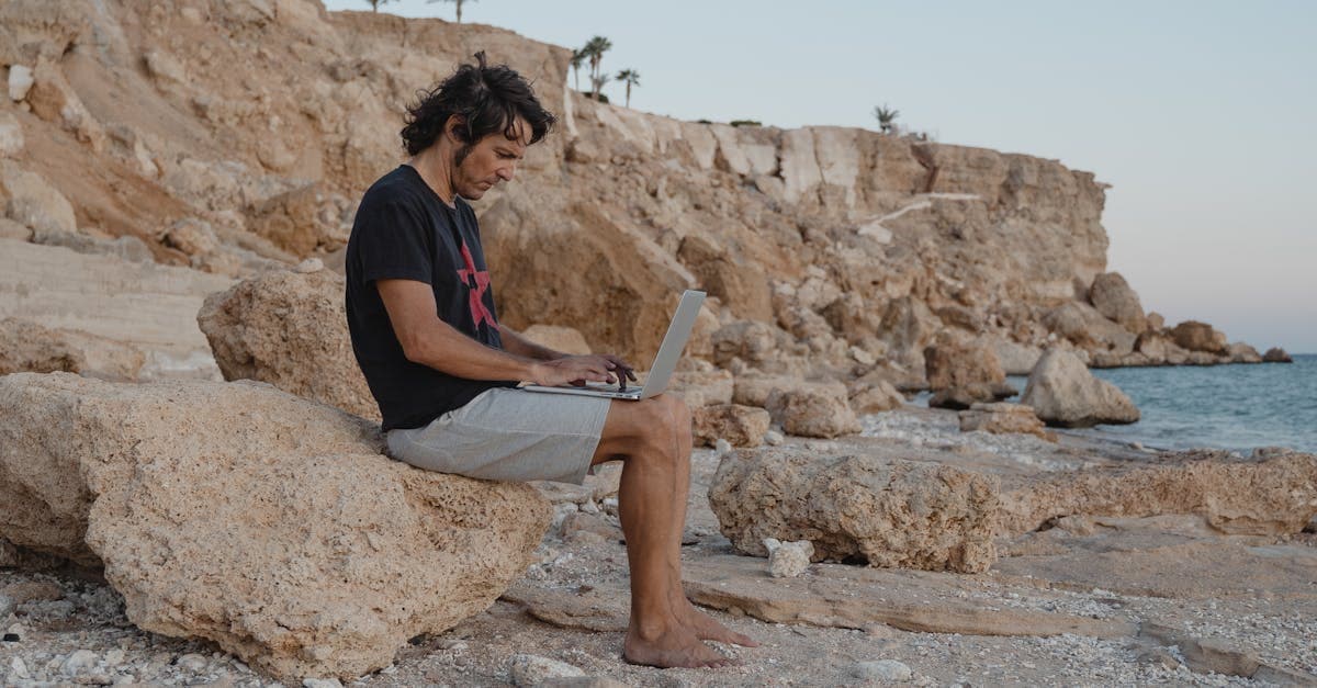 Pria bekerja dengan laptop di bebatuan tepi pantai menikmati sunset sebagai digital travel eSIM profesional