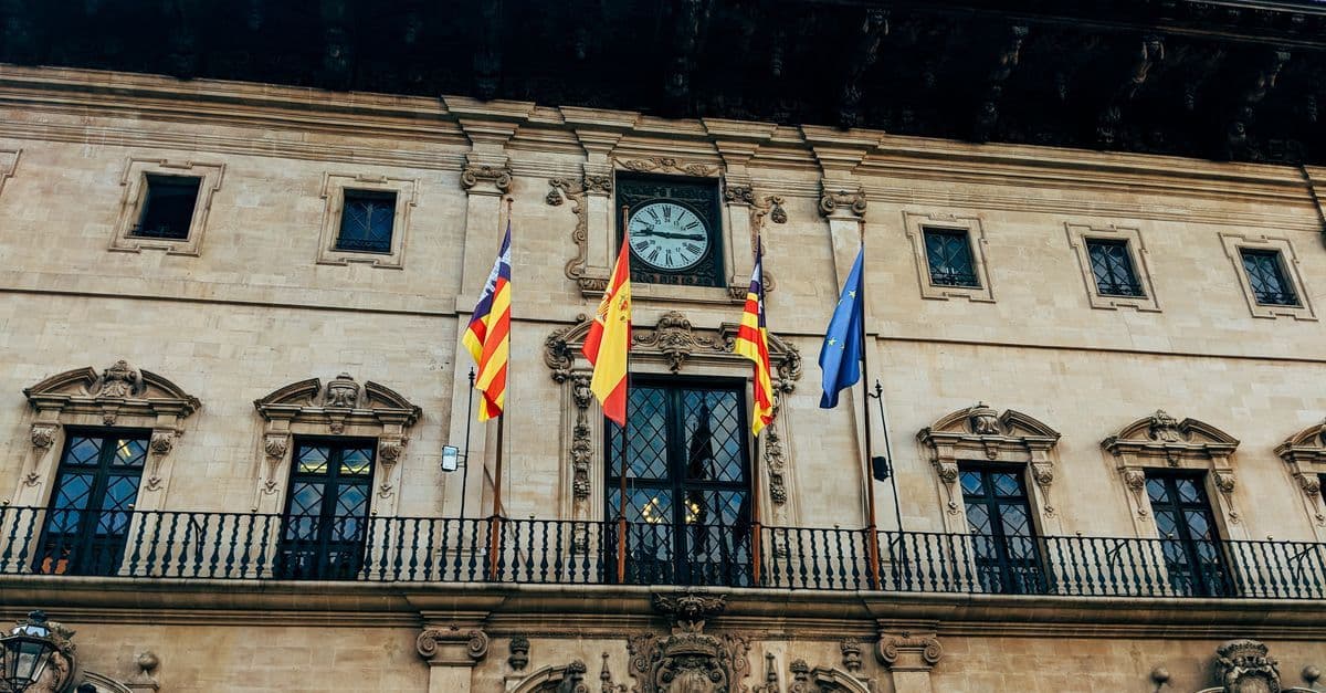 A historic Spanish government building with a clock, balcony and several flags flying above the entrance.