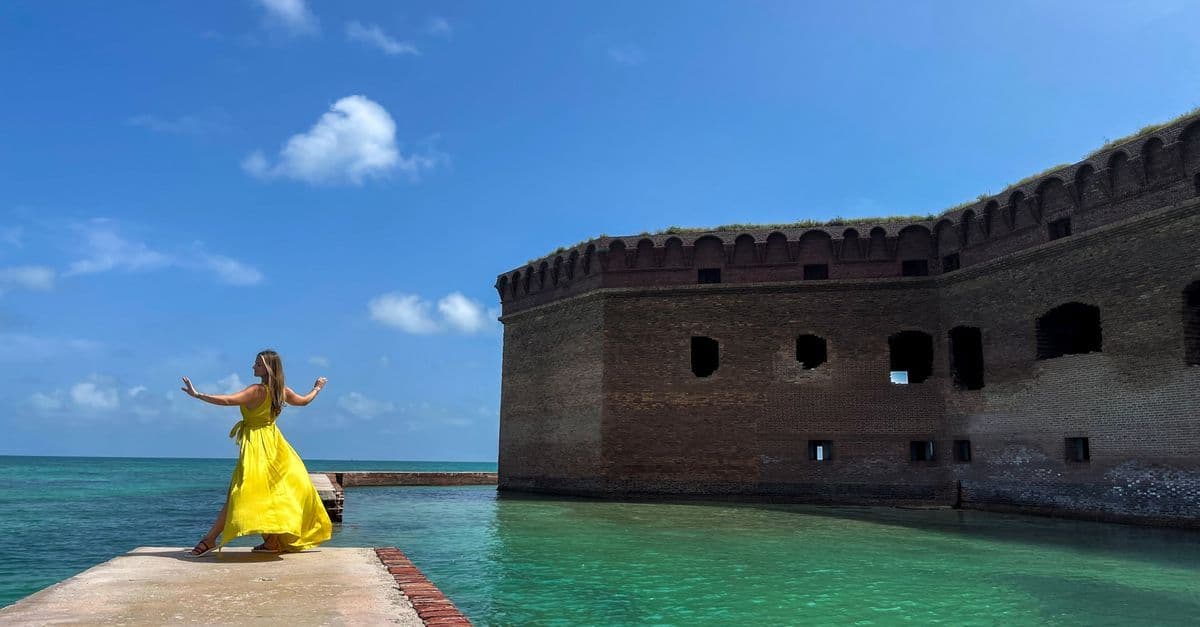 Vrouw in gele jurk poseert op een pier bij een oud fort boven turquoise water onder een strakblauwe lucht.