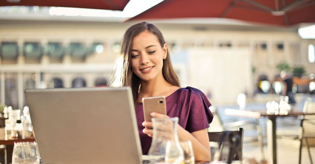 A woman checks her phone beside an open laptop at an outdoor café.
