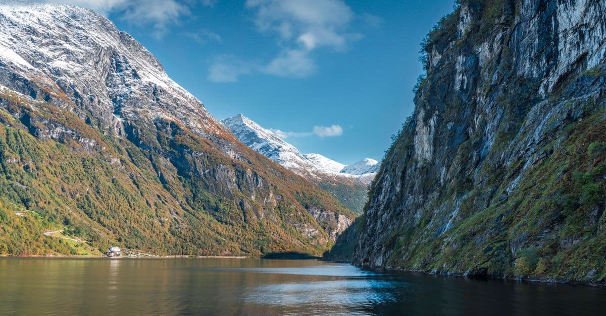 A calm fjord winds between steep mountains, with snow on the peaks and autumn colors along the lower slopes.