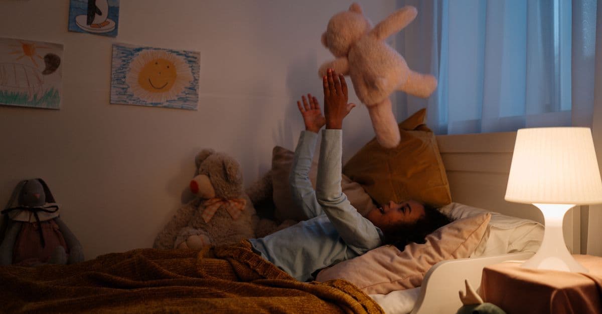 Lachendes Kind liegt im Bett und wirft ein Kuscheltier in die Luft, umgeben von Plüschtieren und Kinderzeichnungen.