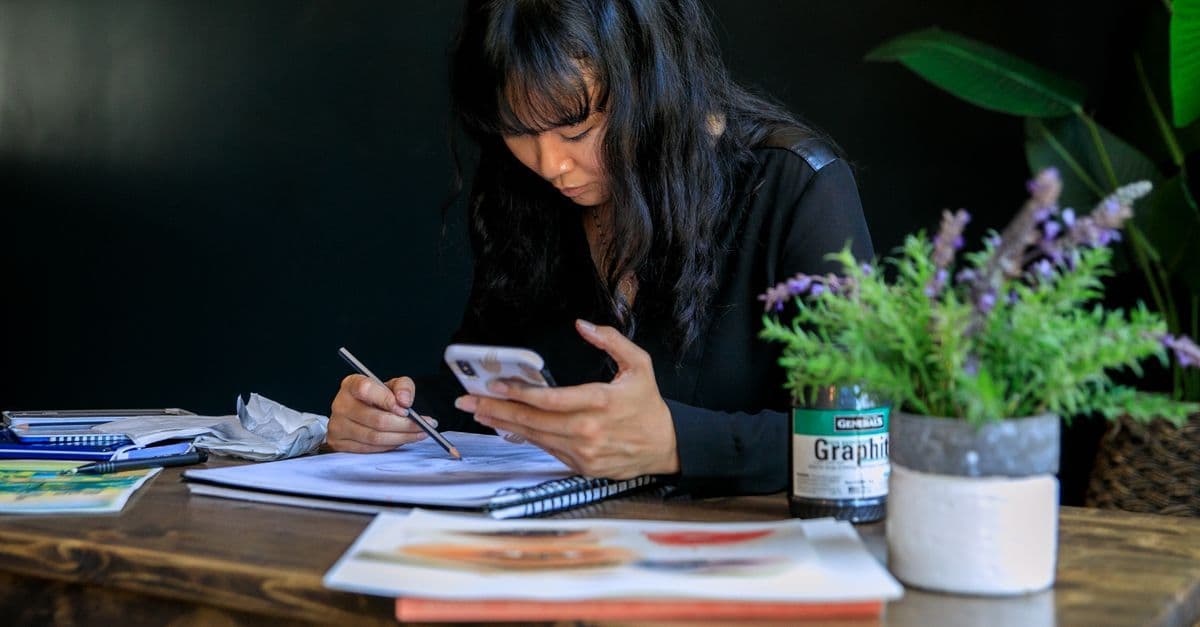 Woman focused on sketching a Gartic Phone drawing prompt while referencing her smartphone screen