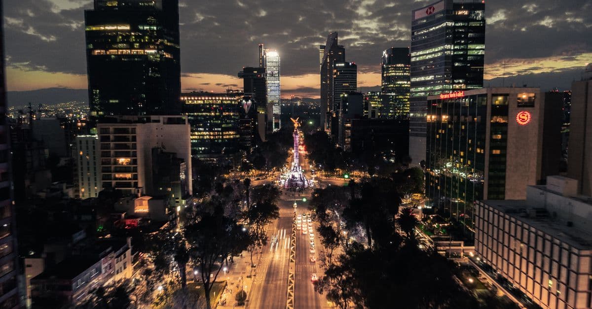 A brightly lit avenue leads toward a monument between skyscrapers beneath a cloudy dusk sky.