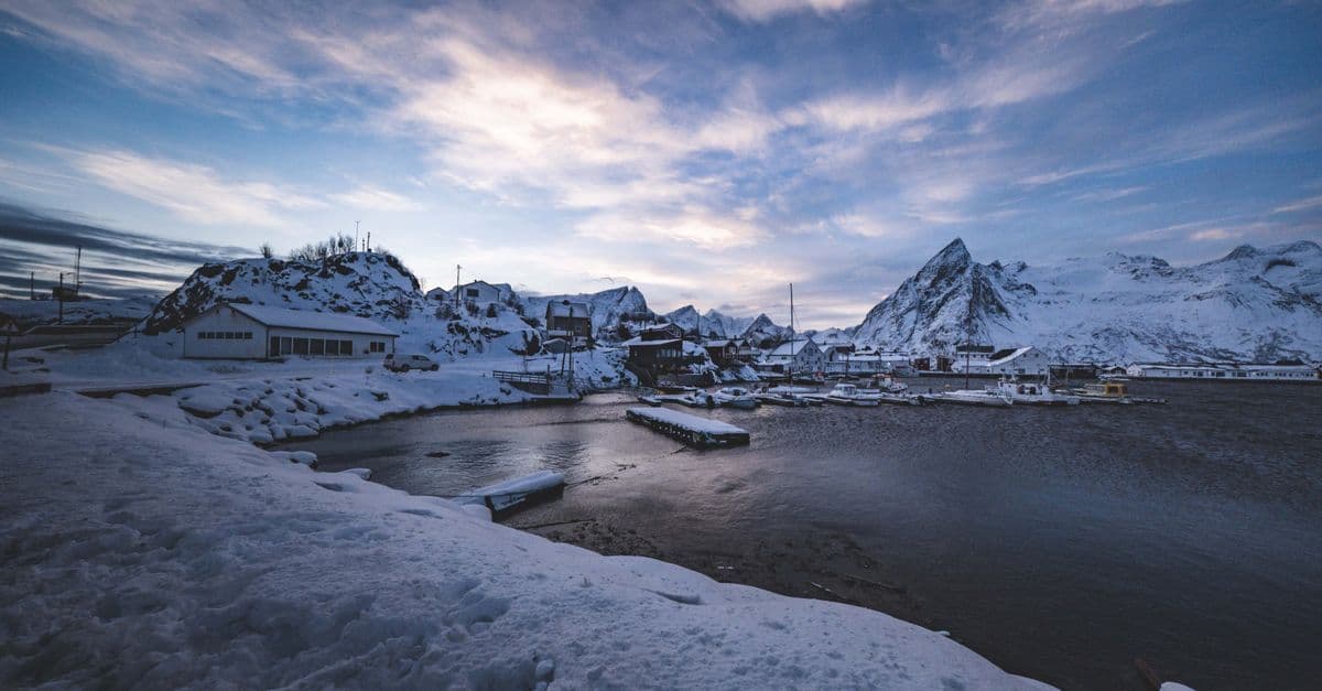 A snow-covered village sits beside a dark harbor, with rugged mountains beneath a cloudy winter sky.