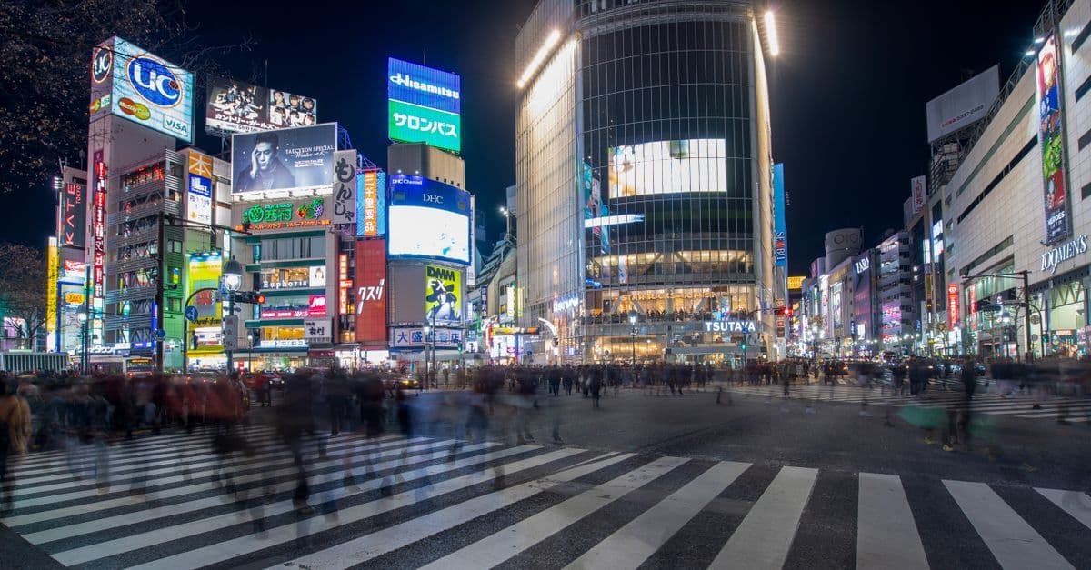Busy nighttime city crosswalk with blurred pedestrians and bright advertising screens illuminating the streets.