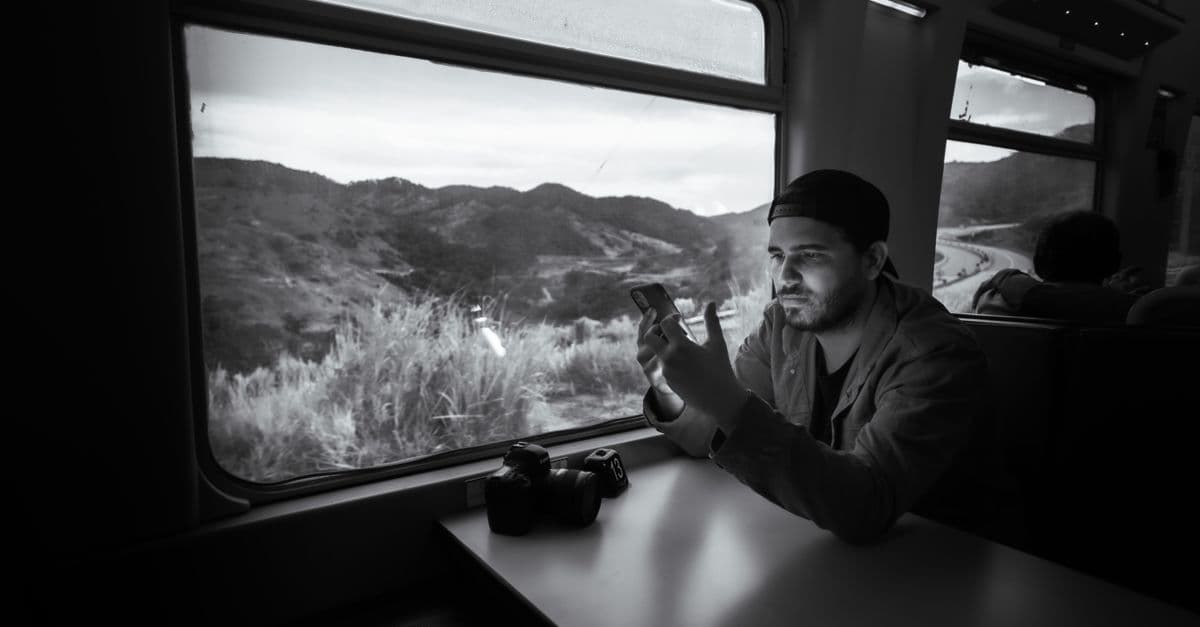 A traveler checks his phone beside a camera on a train, with rugged New Zealand hills visible through the window.