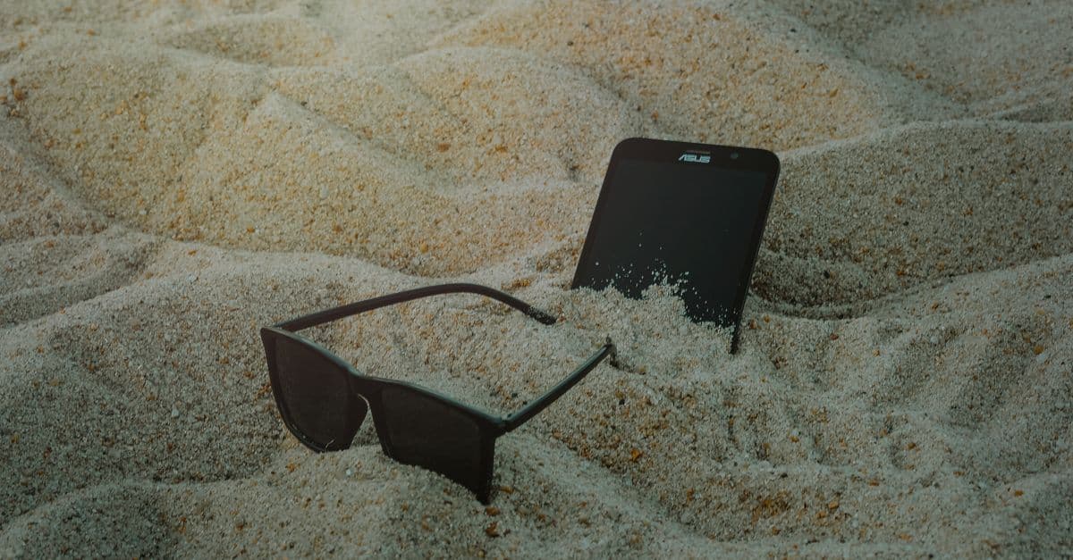 Smartphone e occhiali da sole semisepolti nella sabbia, simbolo del rischio di perdere il telefono in viaggio.