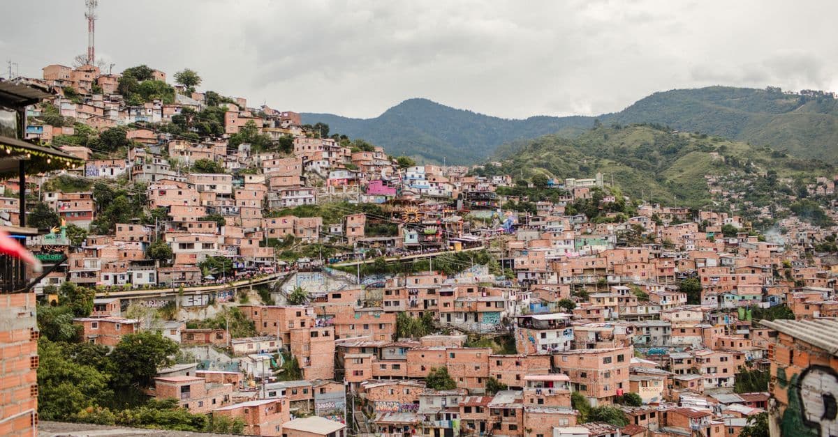 Brick homes climb a hillside in Medellín, Colombia, below green mountains, with a red and white telecom mast on the ridge.