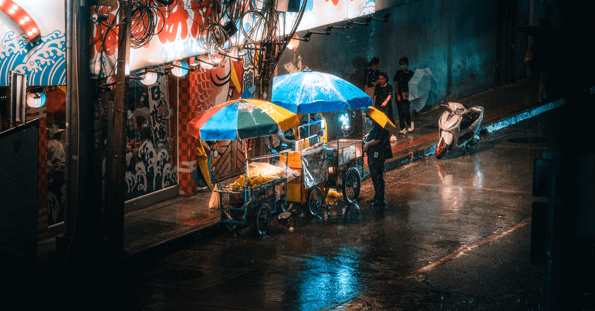 Street food carts shelter under colorful umbrellas as people walk along a rain-soaked street at night.
