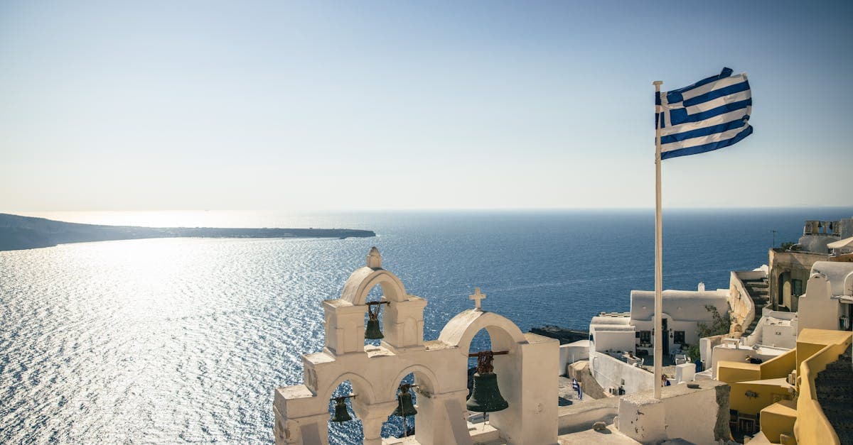 Vita klockorn och grekisk flagga på Santorini med det blå Egeiska havet i bakgrunden.