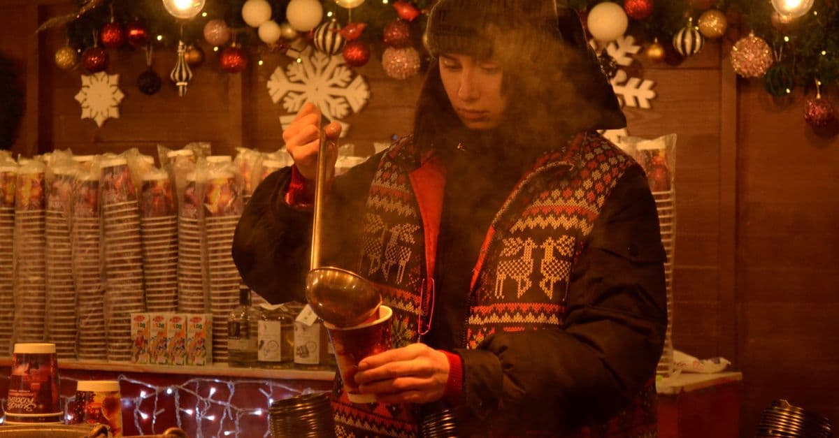 A vendor ladles a hot drink at a warmly lit Christmas market stall decorated with festive ornaments.